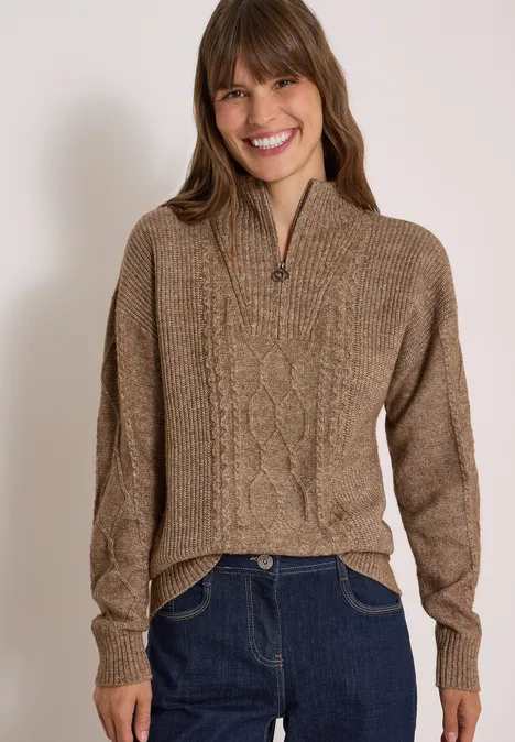 Frau trägt lässigen braunen Zopfmuster-Pullover mit Reißverschluss am Stehkragen, lächelt.