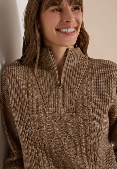 Lächelnde Frau in braunem, grob gestricktem Strickpullover mit Zopfmuster und halbem Reißverschluss.