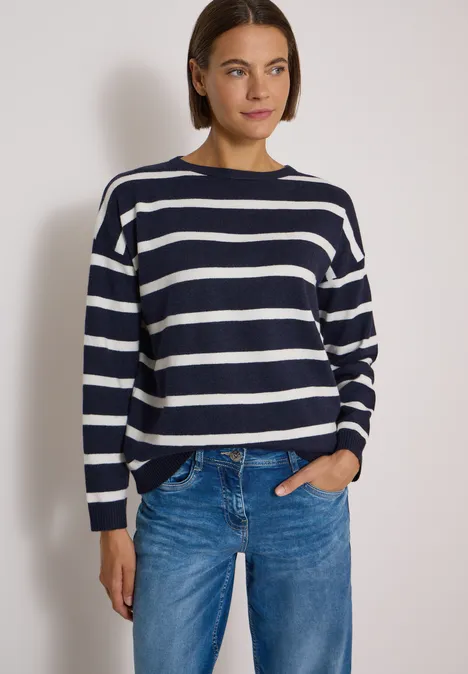 Frau trägt navyweiß gestreiften Pullover und blaue Jeans vor weißem Hintergrund.