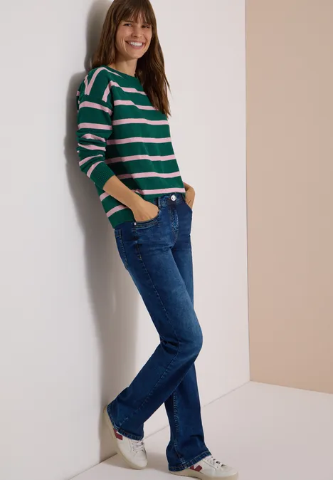 Pullover mit Boat Neck und Streifen dark fir green melange Frau lehnt lächelnd an Wand, trägt gestreiften Pullover, Jeans und Sneaker.