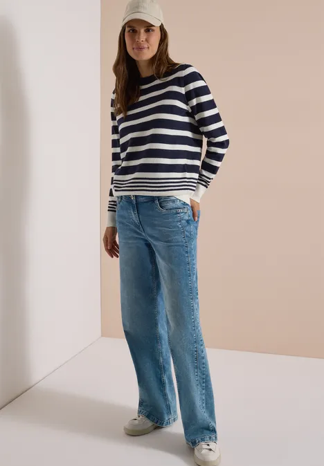 Pullover mit Rundhals und Streifen urban dark blue melange Frau in gestreiftem Pullover, Jeans, Sneakern und Cap steht entspannt mit Händen in den Taschen.