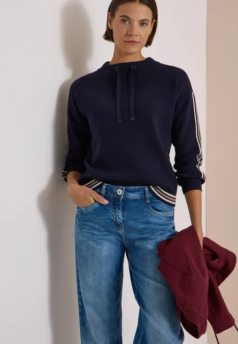 Pullover mit Turtle Neck und College-Details urban dark blue melange Eine Frau in blauem Pullover mit Kordelzug und Jeans hält eine bordeauxrote Jacke.