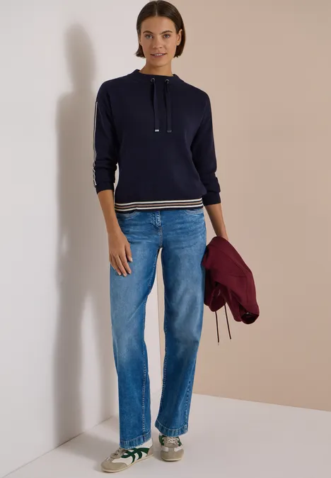 Pullover mit Turtle Neck und College-Details urban dark blue melange Frau in dunkelblauem Hoodie, Jeans und Sneakern hält eine burgunderrote Jacke.