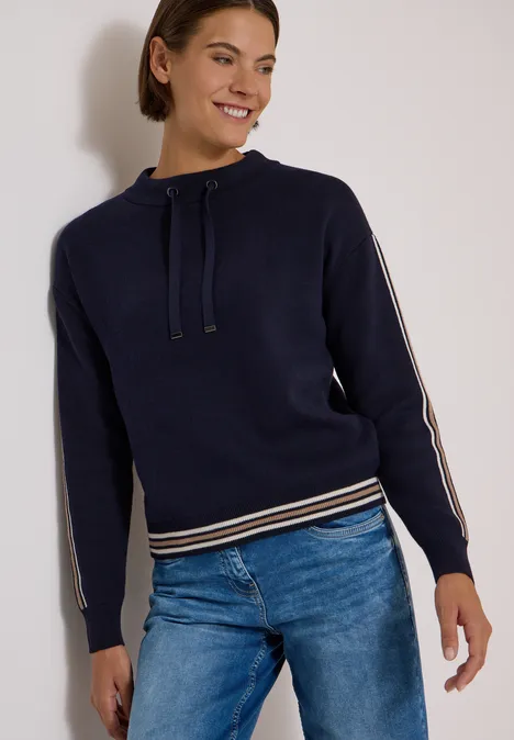 Pullover mit Turtle Neck und College-Details urban dark blue melange Frau in blauem Kapuzenpullover mit Streifen an Ärmeln und Bund, kombiniert mit Jeans, steht lächelnd.