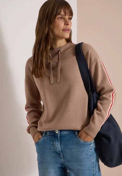 Trui met col en college-details creamy mocha melange Vrouw in taupekleurige capuchontrui met gestreepte mouwen, spijkerbroek en donkerblauwe tas.