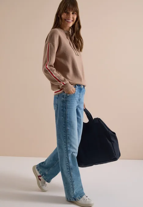 Trui met col en college-details creamy mocha melange Vrouw in beige trui, blauwe jeans en sneakers, draagt een grote blauwe tas, glimlachend voor een effen achtergrond.