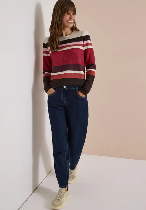 Sweter z okrągłym dekoltem i w paski rhubarb melange Uśmiechnięta kobieta opiera się o jasną ścianę, ma na sobie pasiowany sweter, luźne dżinsy i białe adidasy.