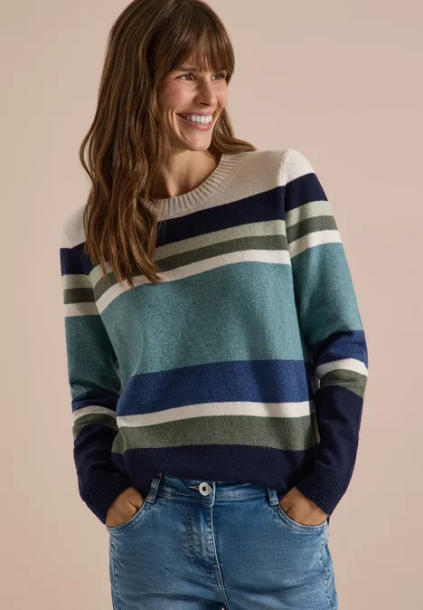Sweter z okrągłym dekoltem i w paski mineral blue melange Uśmiechnięta kobieta w pasiastym swetrze w odcieniach niebieskiego, zielonego i białego, z dżinsami, na beżowym tle.