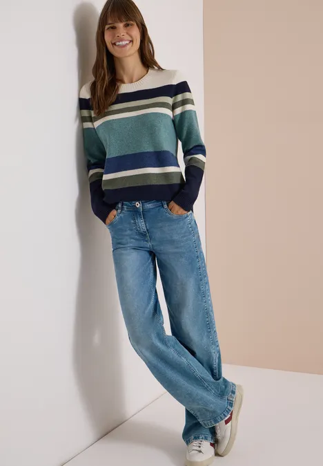 Sweter z okrągłym dekoltem i w paski mineral blue melange Uśmiechnięta kobieta opiera się o ścianę, ma na sobie pasiasty sweter, niebieskie dżinsy i trampki.