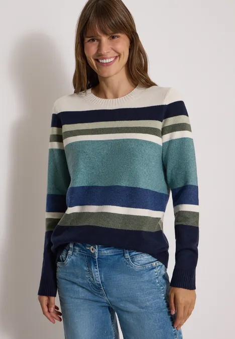 Sweter z okrągłym dekoltem i w paski mineral blue melange Uśmiechnięta kobieta w pasiastym swetrze w odcieniach niebieskiego, zielonego i białego oraz w dżinsach na jasnym tle.