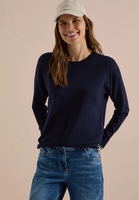 Pullover mit Rundhals und Layering-Detail urban dark blue Frau mit weißer Baseballmütze, blauem Pullover und Jeans lächelt locker in die Kamera.