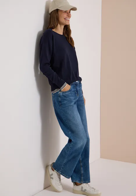 Pullover mit Rundhals und Layering-Detail urban dark blue Frau lehnt lächelnd in Jeans, dunkelblauem Pullover und Cap an heller Wand.