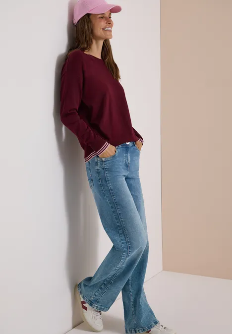 Pullover mit Rundhals und Layering-Detail red wine Eine lächelnde junge Frau in Jeans, burgunderfarbenem Pullover, rosa Cap und Sneakern lehnt entspannt an der Wand.