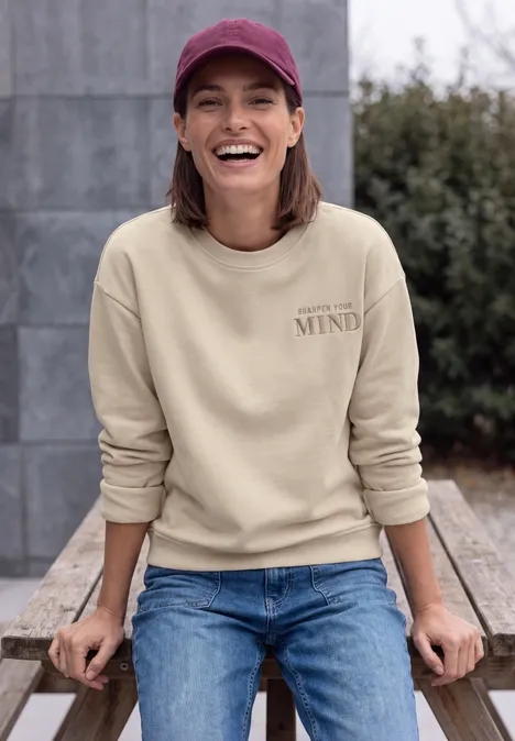 Eine lächelnde Frau in Jeans und beigem Sweatshirt mit Cap sitzt auf einer Holzbank vor einer grauen Wand.