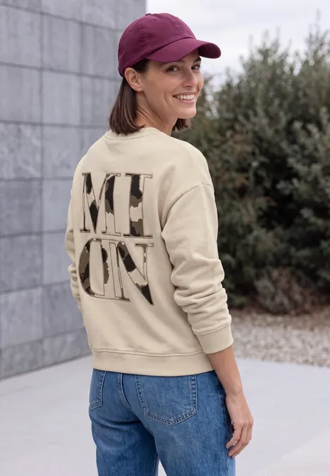 Eine Frau in Jeans und beigem Pullover mit großem Schriftzug auf dem Rücken steht draußen und lächelt zur Kamera.