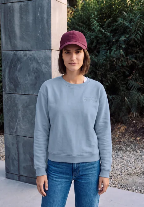 Sweatshirt mit Rundhals und Backprint cosmopolitan blue Frau steht vor Steinwand im Garten, trägt hellblaues Sweatshirt, Jeans und rotes Cap.