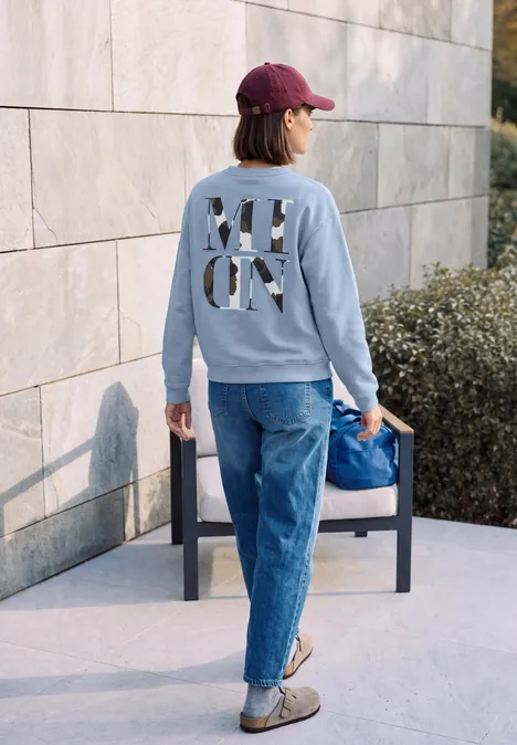 Sweatshirt mit Rundhals und Backprint cosmopolitan blue Rückansicht einer Person in roter Mütze, grauem Pullover mit Schriftzug, blauen Jeans und blauer Tasche.