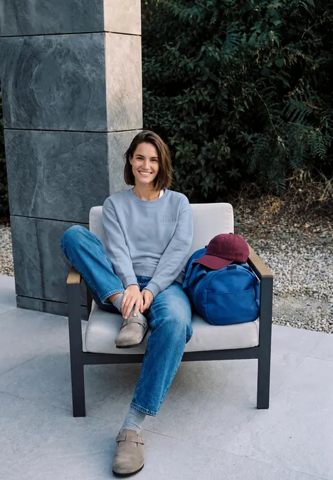 Sweatshirt mit Rundhals und Backprint cosmopolitan blue Junge Frau in Jeans und Pullover sitzt auf modernem Stuhl im Freien, lächelt entspannt.