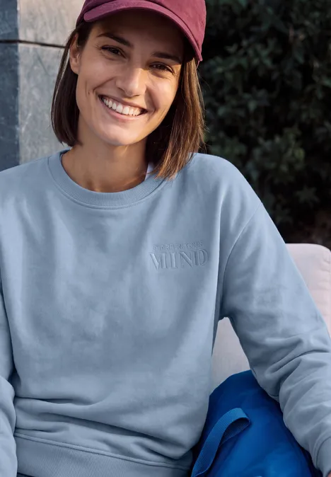 Sweatshirt mit Rundhals und Backprint cosmopolitan blue Junge Frau mit roter Baseballkappe und hellem Sweatshirt sitzt lächelnd vor Grünpflanzen.