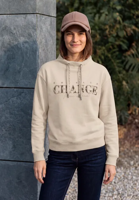 Junge Frau vor Steinmauer trägt hellen Hoodie mit Schriftzug, Jeans und Cap, steht lächelnd im Freien.
