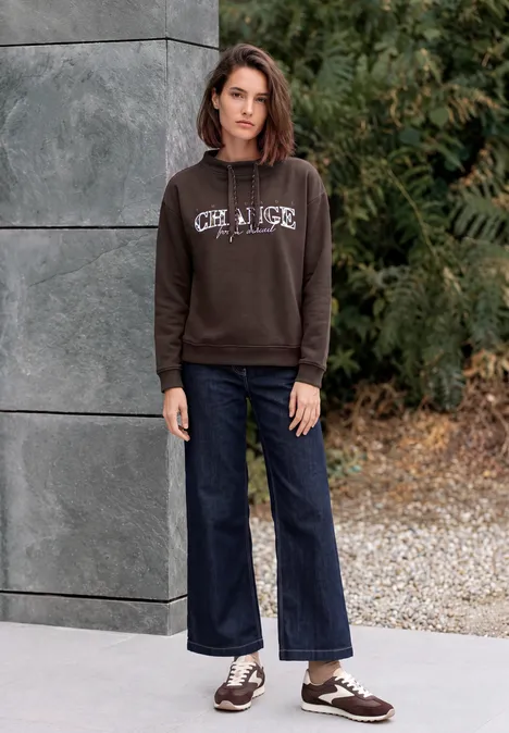 Frau im braunen Kapuzenpullover und weiten Jeans steht vor einer Mauer und grünem Strauchwerk.