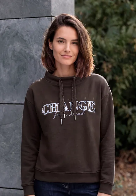 Eine lächelnde Frau im braunen Hoodie steht vor einer Steinwand und grünen Sträuchern.