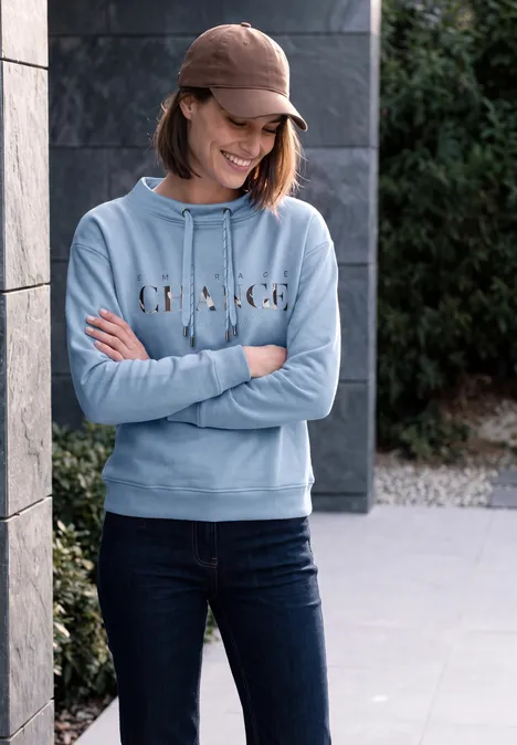 Frau in lässigem Outfit: blaue Kapuzenjacke, Jeans und Cap lehnt lächelnd an einer Wand.