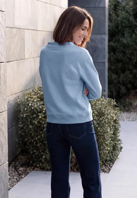 Frau in blauem Pullover und Jeans lehnt seitlich an Hauswand, Heckengebüsch im Hintergrund.