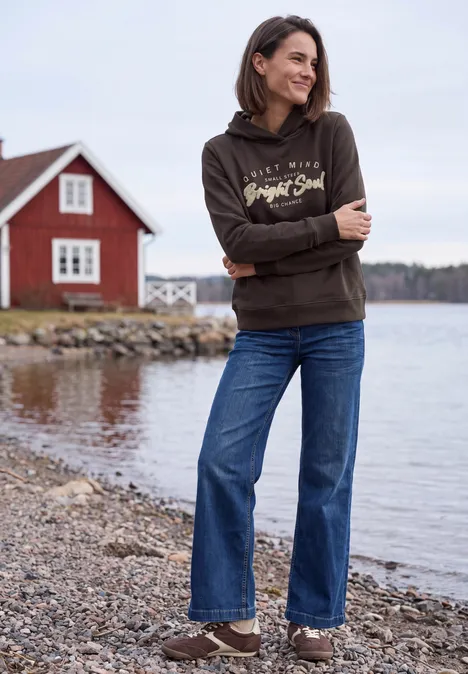 Frau in Hoodie und Jeans steht lächelnd am Ufer eines Sees mit rotem Häuschen im Hintergrund.