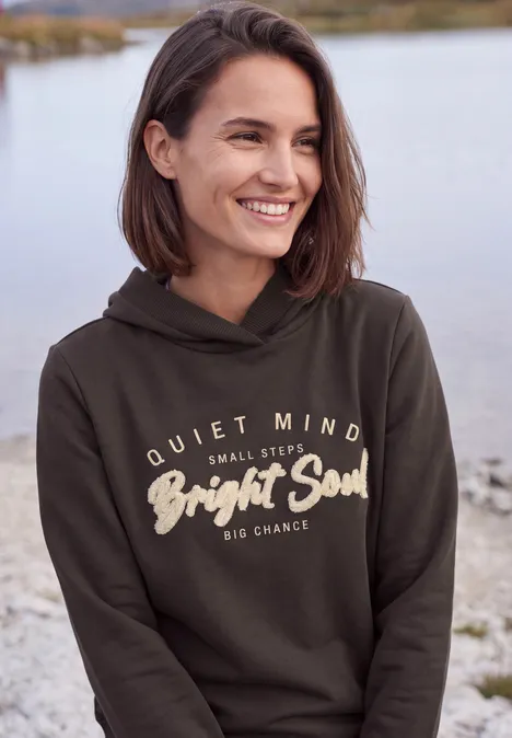 Lächelnde Frau in Kapuzenpullover mit Schriftzug am Ufer eines Sees.
