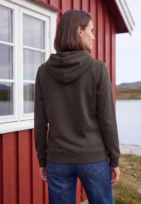 Rückansicht einer Person in Kapuzenpullover und Jeans vor einem roten Haus nahe dem Wasser.