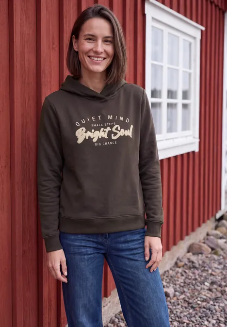 Eine lächelnde Frau steht vor einem roten Holzhaus, trägt Jeans und einen Hoodie mit Aufschrift 'Bright Soul'.