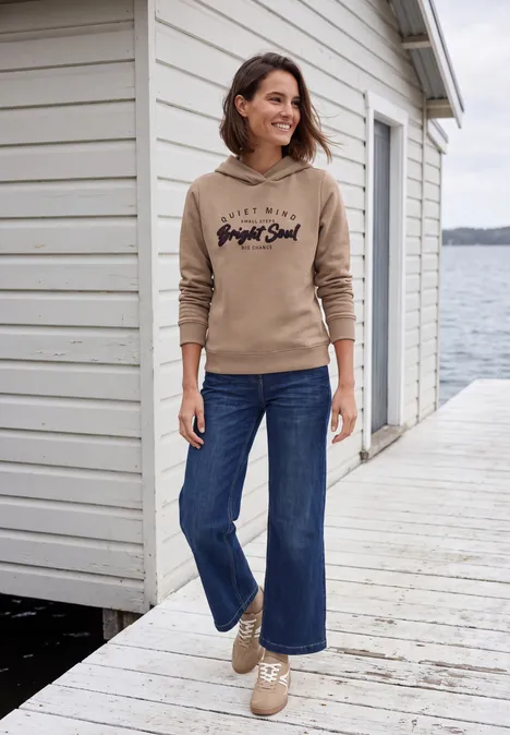 Femme en sweat à capuche beige et jean, souriante sur un ponton devant une maison au bord de l’eau.