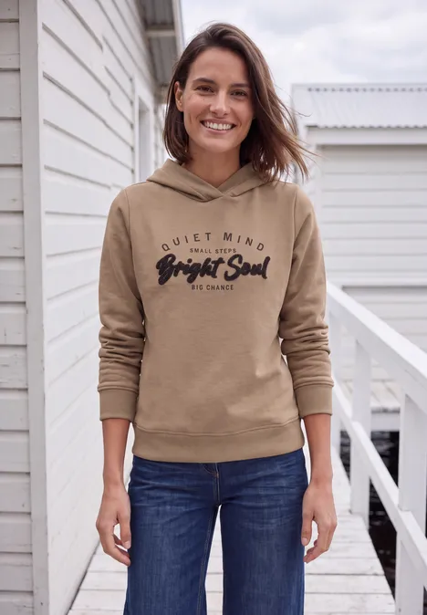 Femme souriante en sweat à capuche beige et jean, debout sur une terrasse.