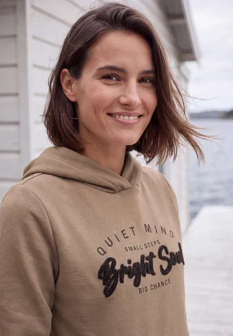 Femme souriante en sweat à capuche marron, debout près de l'eau devant une maison en bois.