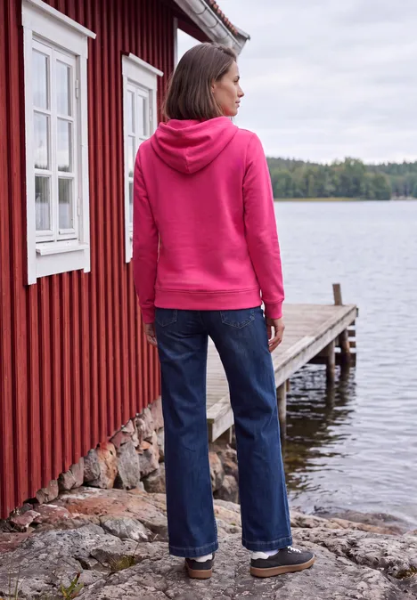 Frau in pinkem Kapuzenpullover und Jeans steht am Ufer neben roter Holzhütte und blickt auf den See.