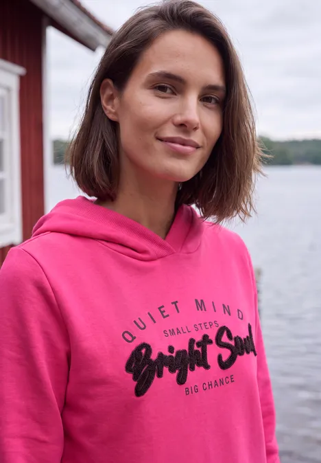Junge Frau in pinkem Hoodie steht lächelnd am Seeufer vor rotem Holzhaus.