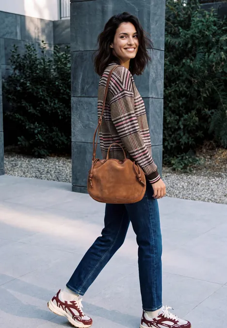 Sweat-shirt à col rond et motif à carreaux dark toffee brown Eine lächelnde Frau in Jeans, kariertem Pullover und Sneakers trägt eine braune Tasche vor modernem Gebäude.