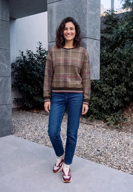 Sweat-shirt à col rond et motif à carreaux dark toffee brown Eine lächelnde Frau trägt Pullover und Jeans und steht vor einer Mauer mit Pflanzen.