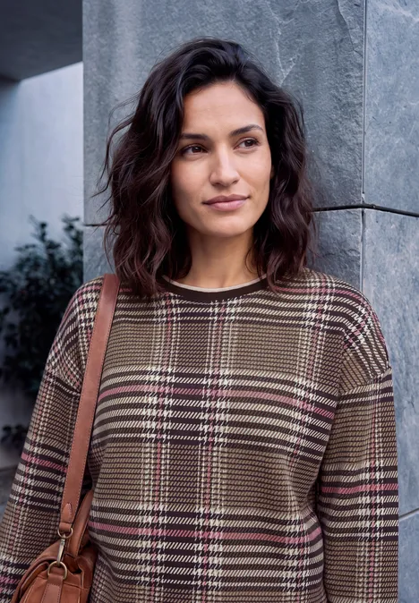 Sweat-shirt à col rond et motif à carreaux dark toffee brown Junge Frau mit gelocktem Haar in kariertem Oberteil lehnt an grauer Säule.
