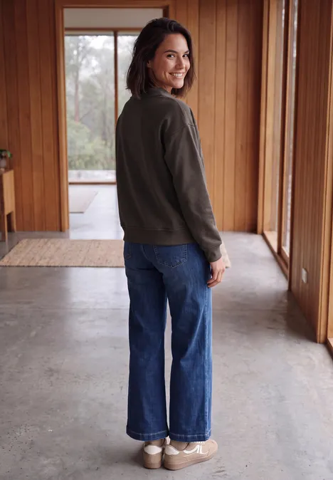 Une femme en veste verte et jeans bleu sourit en se retournant dans un couloir lumineux en bois.