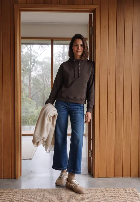Femme en jean et sweat à capuche, tenant une veste, debout dans une porte en bois.
