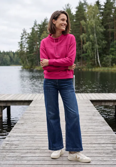 Een vrouw in een roze capuchontrui en spijkerbroek lacht en staat op een houten steiger bij een meer met bos op de achtergrond.