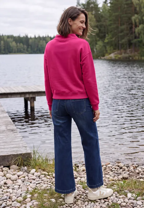 Vrouw in een roze hoodie, blauwe jeans en witte schoenen staat bij een meer met bos op de achtergrond.
