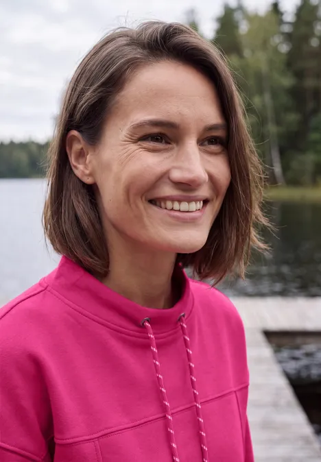 Een lachende vrouw met kort bruin haar draagt een roze trui bij een rustig meer.