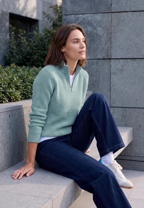 Pull uni à fermeture zip multi lapis melange Femme en tenue décontractée assise sur un banc en pierre, regard détendu vers l'horizon.