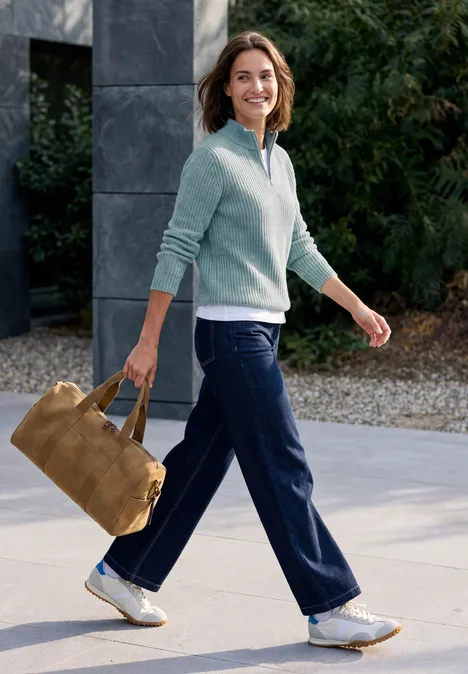 Pull uni à fermeture zip multi lapis melange Femme souriante marchant dehors, porte un pull bleu clair, un pantalon bleu et un sac en daim marron.