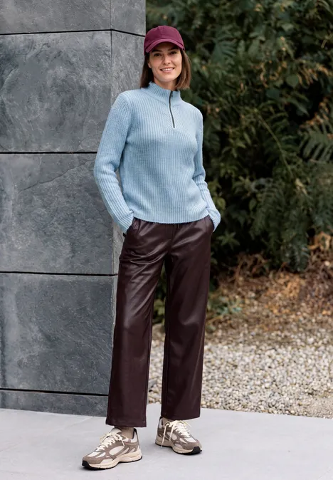 Eine Frau in blauem Pullover, roter Cap und Lederhose lehnt lächelnd an einer Mauer im Freien.