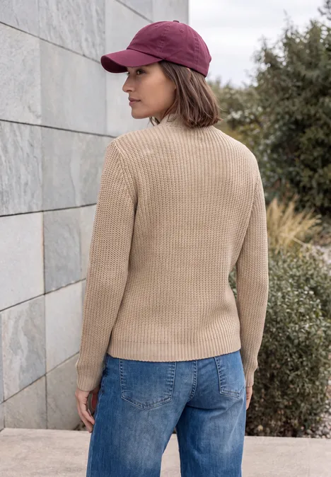 Pull uni à fermeture zip creamy mocha melange Femme de profil devant un mur en pierre, casquette rouge, pull beige et jean bleu.