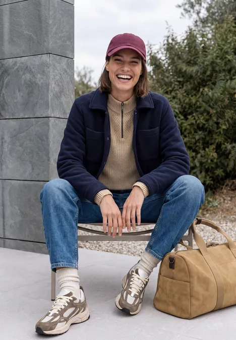 Pull uni à fermeture zip creamy mocha melange Une jeune femme assise sur un banc en plein air, vêtue de style décontracté, casquette et souriante.